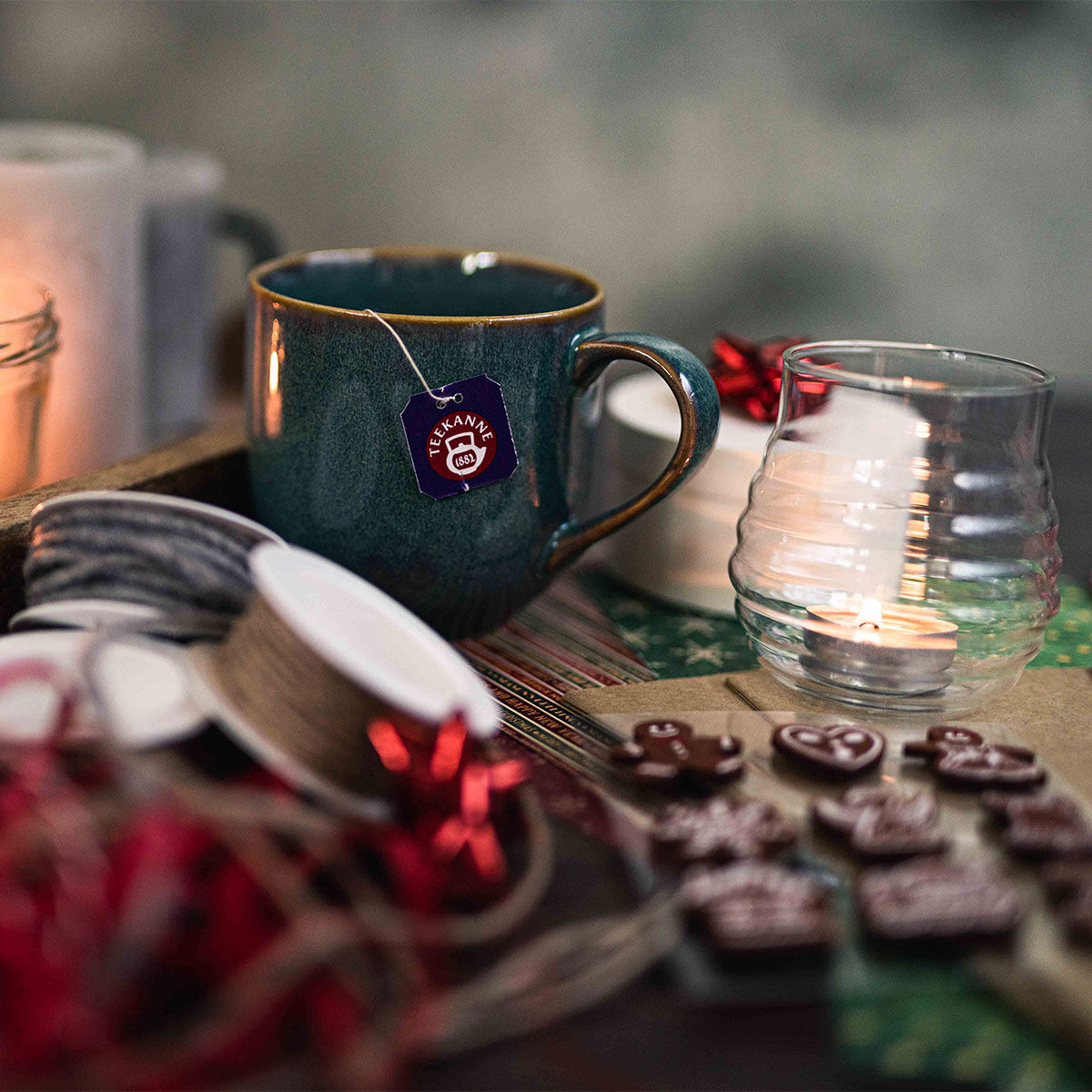 Eine blaugrüne Tasse Sternenleuchten-Tee mit einem Teebeutel, einer brennenden Kerze in einem Glashalter, Bandrollen und Schokoladenlebkuchenfiguren stehen auf einem festlich gedeckten Tisch und schaffen eine gemütliche und einladende Atmosphäre.