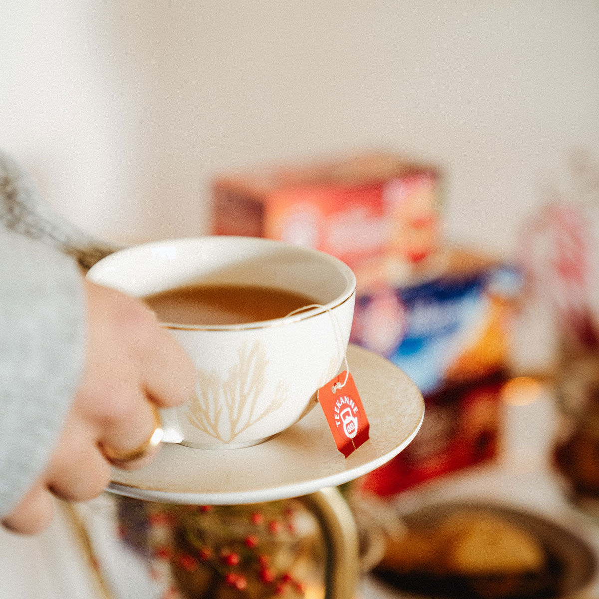 Eine Person hält eine weiße Teetasse mit dem roten Etikett „Süßer Bratapfel“ auf einer passenden Untertasse. Im Hintergrund sind verschwommene Schachteln und Snacks zu sehen.