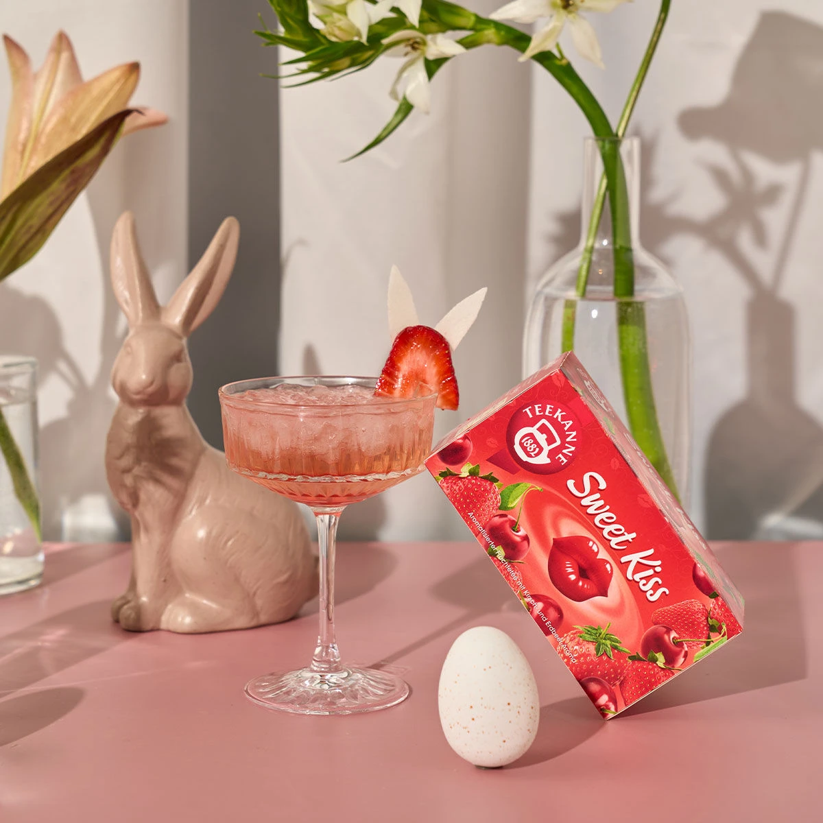 Ein rosa Cocktail mit Erdbeergarnitur steht neben einem Keramikhasen, einem gesprenkelten Ei, weißen Blumen in einer Vase und einer roten Schachtel Sweet Kiss auf einem rosa Tisch.