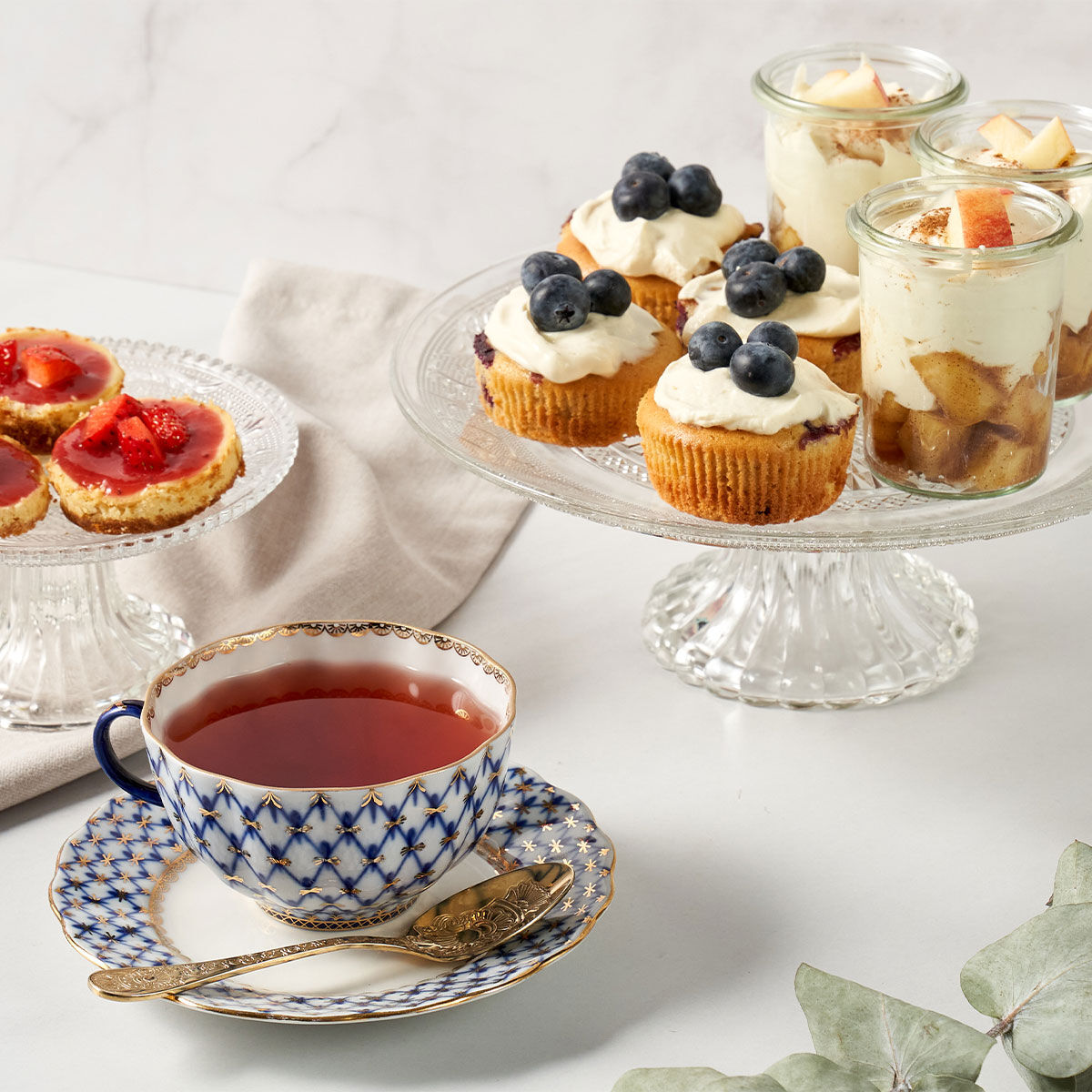 Eine Tasse Tee von Sweet Teatime steht in einer dekorativen Teetasse vor einem Glasständer, auf dem Blaubeer-Cupcakes, Apfelparfait-Gläser und Erdbeertörtchen auf einer Marmoroberfläche mit einer Stoffserviette stehen.