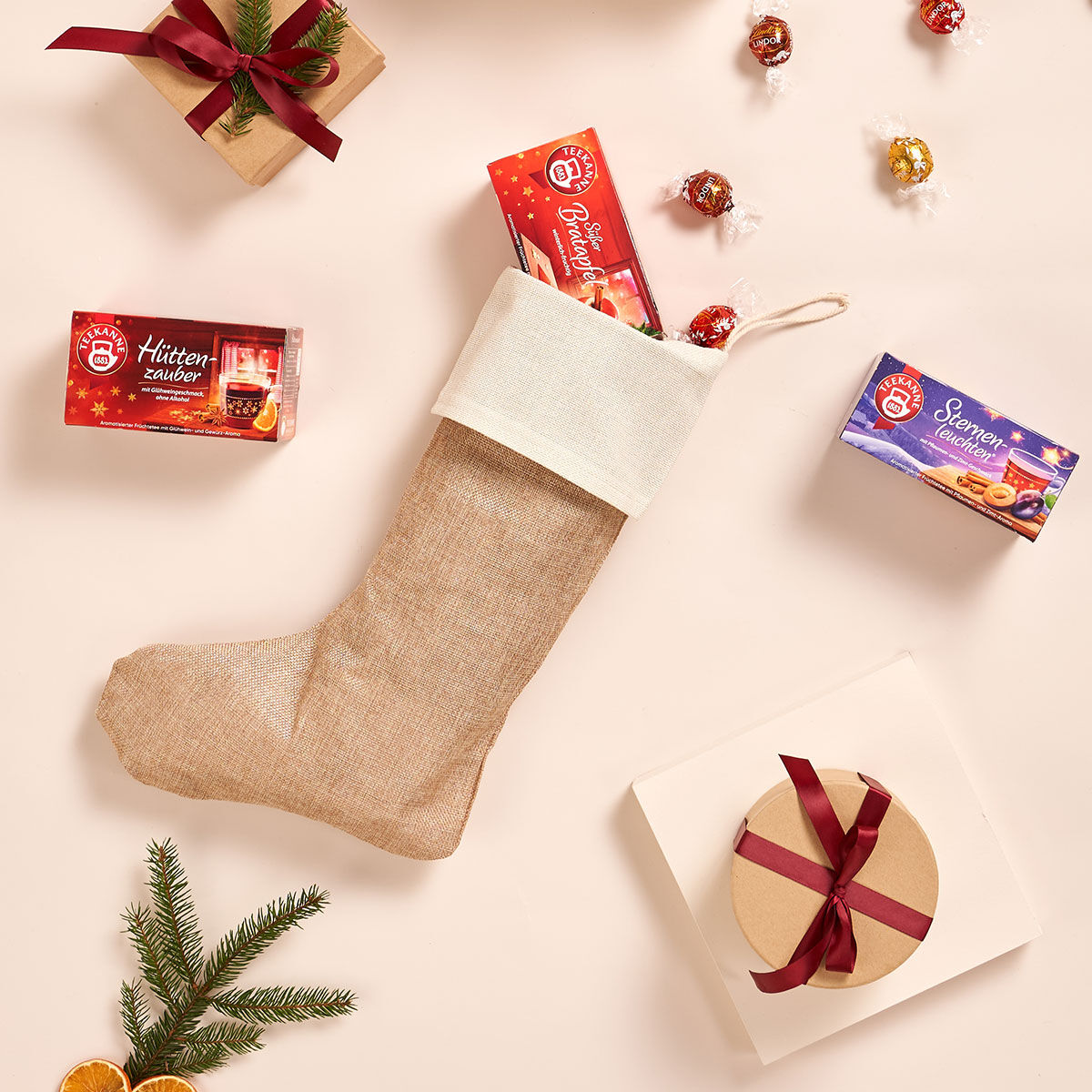 Das Winter Wonderland Geschenkset – ein Weihnachtsstrumpf aus Sackleinen, gefüllt mit Pralinenschachteln – liegt zwischen verpackten Geschenken, Pralinen und immergrünen Zweigen und zaubert auf einem hellen Hintergrund eine gemütliche Weihnachtsmagie.