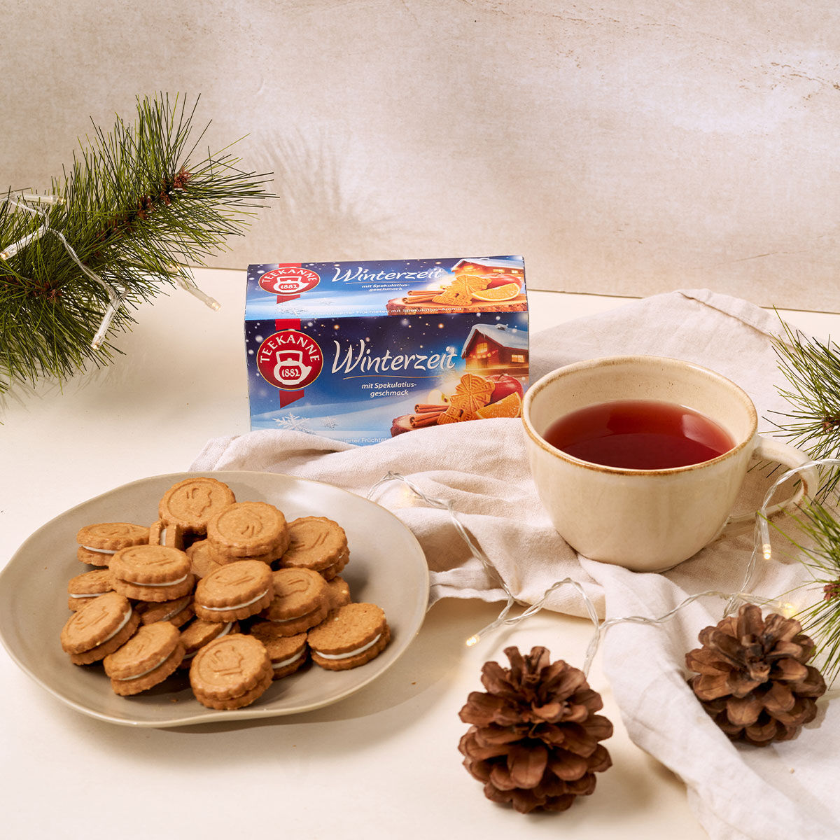 Ein Teller mit Spekulatius und Wintertee steht mit Tannenzweigen, einem Tannenzapfen und einer Winterzeit-Dose auf einem Tisch. Eine leichte Tischdecke verleiht der Szene eine gemütliche, festliche Atmosphäre.