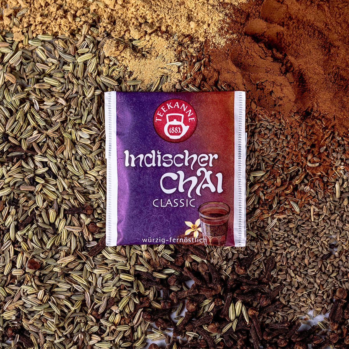 Eine Packung Indischer Chai Classic Tee liegt auf einer Oberfläche, die mit würzigen Gewürzen wie Zimt, Fenchelsamen, Nelken und Anissamen bestreut ist.