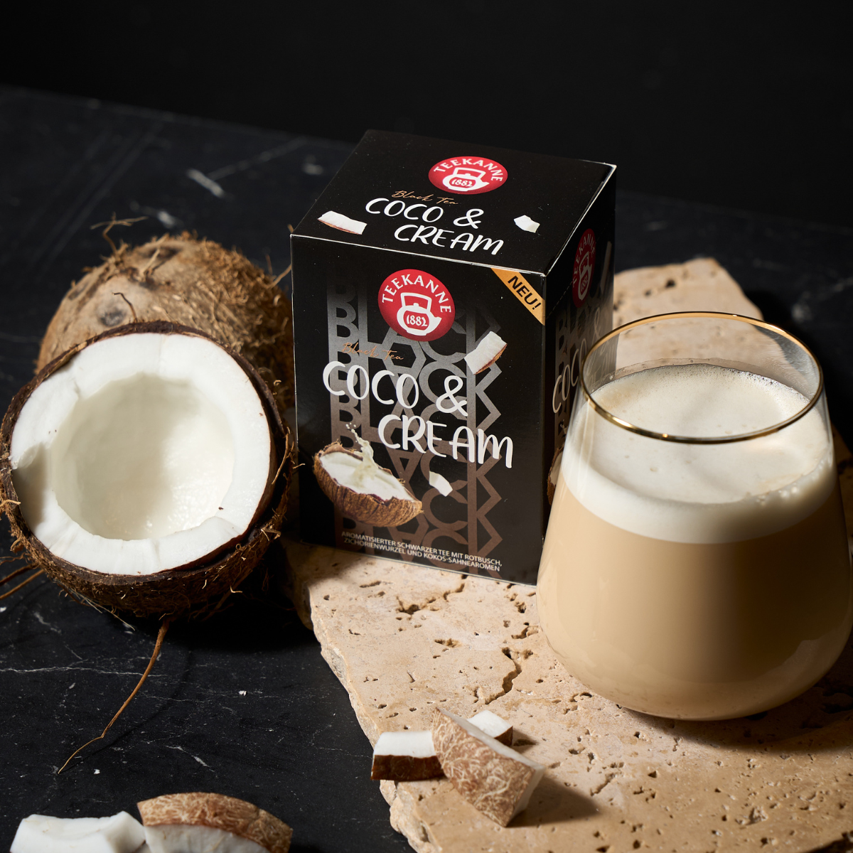 Ein Glas cremiges Kokosnussgetränk steht neben einer halbierten Kokosnuss, Kokosnussstücken und einer Schachtel mit der Aufschrift „Black Tea Coco & Cream“ auf einer strukturierten beigen Oberfläche mit dunklem Hintergrund.