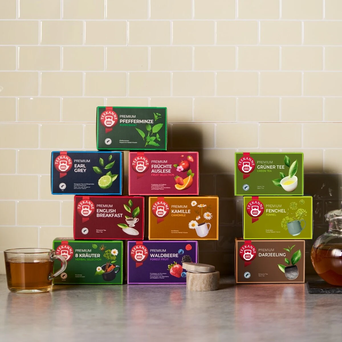 Zehn farbenfrohe Kisten mit Premium Früchte Auslese-Tee stehen ordentlich in zwei Reihen auf einer Theke, begleitet von zwei Tassen Tee und einer gläsernen Teekanne, die vor einer beigefarbenen Kachelwand stehen.