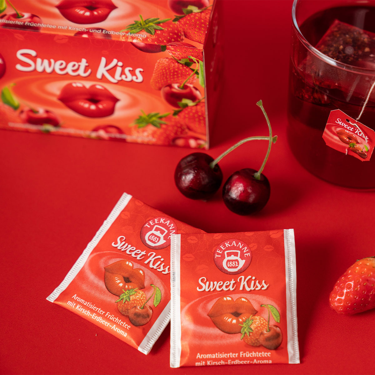 Zwei Verführerische Früchtetees Mix & Match Teebeutel, Kirschen, eine Erdbeere, ein Glas Tee und die mit Lippen- und Beerenmotiven verzierte Schachtel sind auf einer roten Fläche arrangiert und sorgen für eine unwiderstehlich verführerische Präsentation.