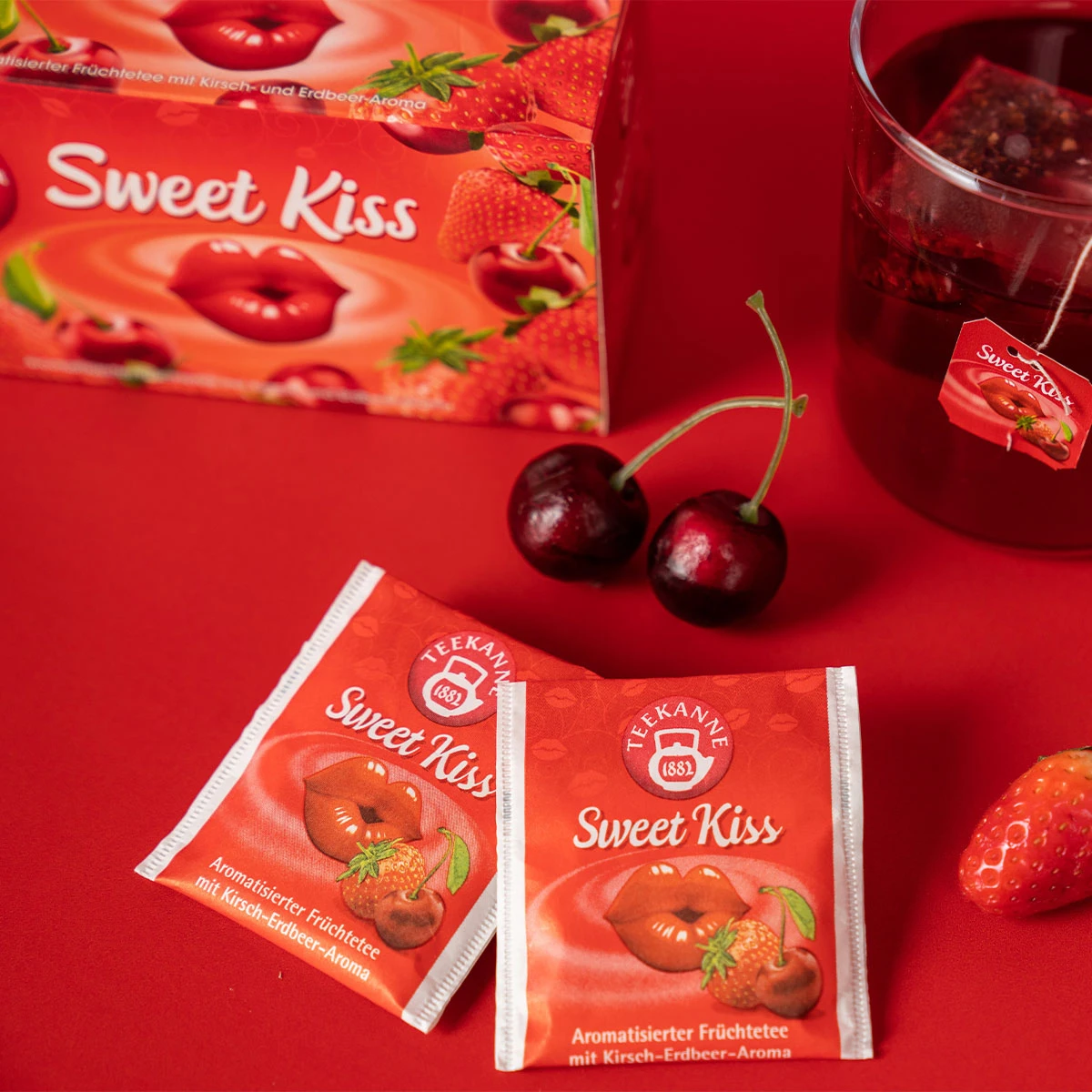 Zwei Verführerische Früchtetees Mix & Match Teebeutel, Kirschen, eine Erdbeere, ein Glas Tee und die mit Lippen- und Beerenmotiven verzierte Schachtel sind auf einer roten Fläche arrangiert und sorgen für eine unwiderstehlich verführerische Präsentation.
