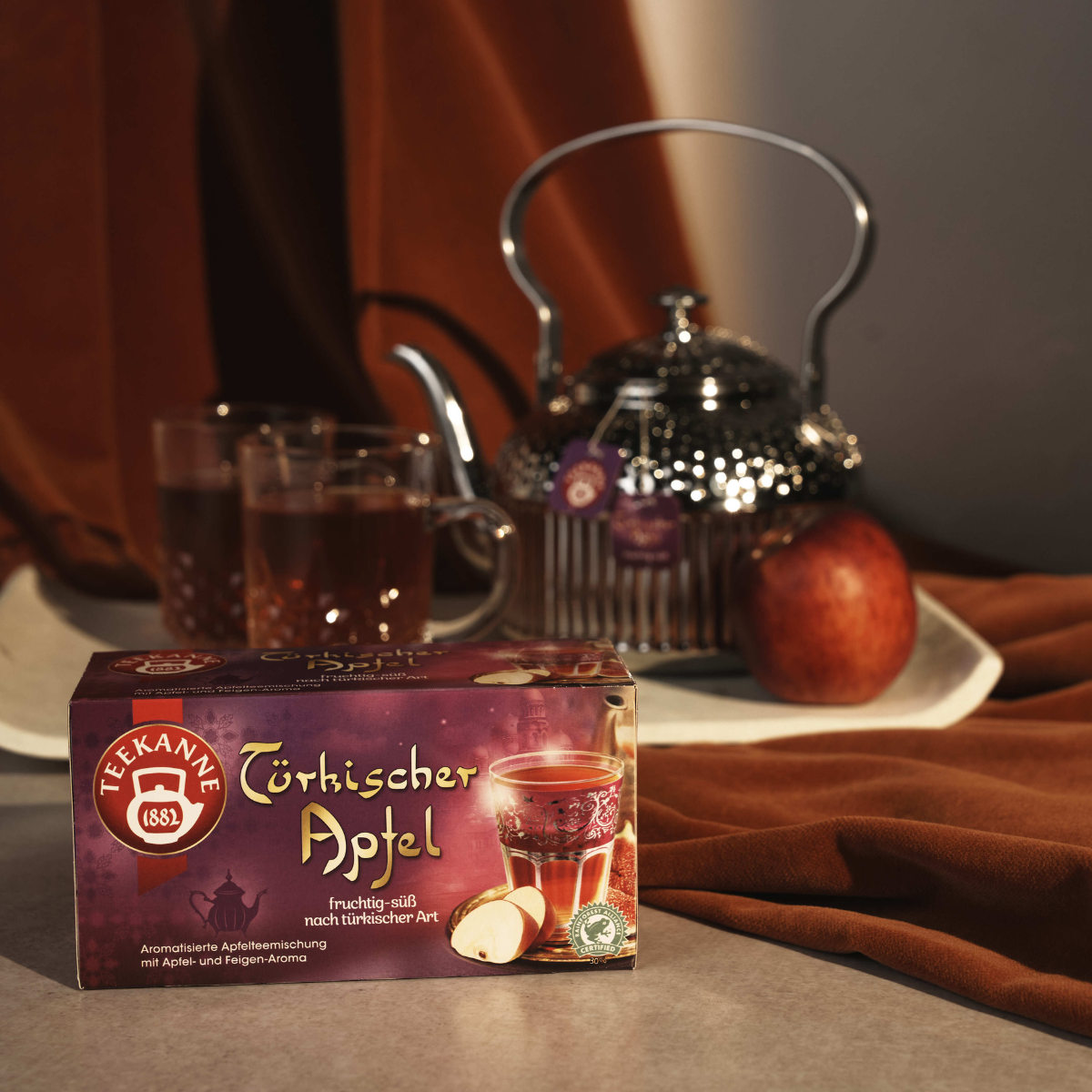 Eine Schachtel Türkischer Apfeltee steht auf einem Tisch neben einem Glas Tee, einer Teekanne und einem Apfel, alles in warmes Licht getaucht, das an die wohltuenden Aromen der Türkei erinnert.