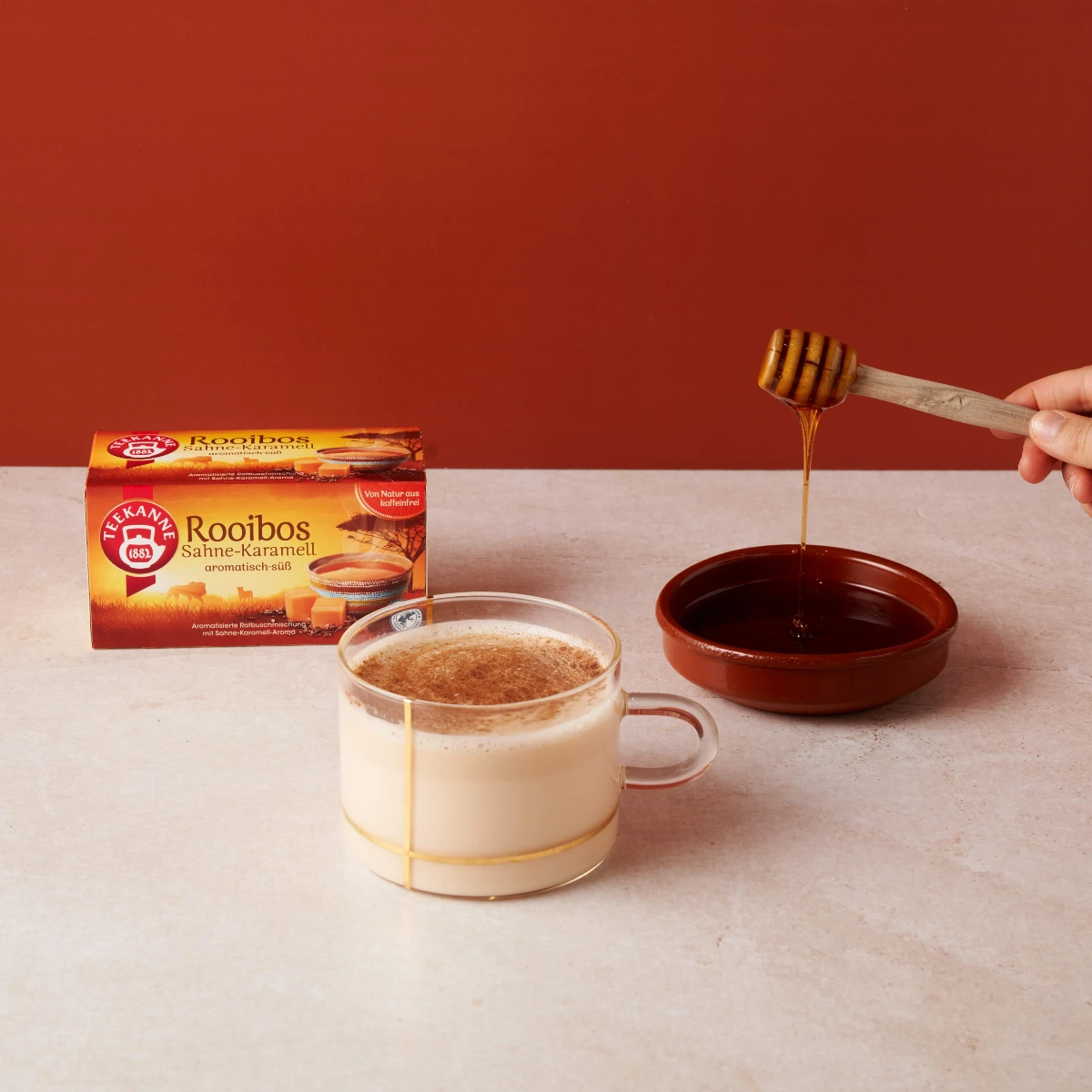 Eine Glastasse Rooibos Sahne-Karamell mit Milch steht auf einer hellen Ablage, während Honig in eine Schale geträufelt wird. Hinter der Tasse befindet sich die Rooibos Sahne-Karamell-Box, die cremige Karamell- und Rooibos-Aromen bei jedem Schluck verspric