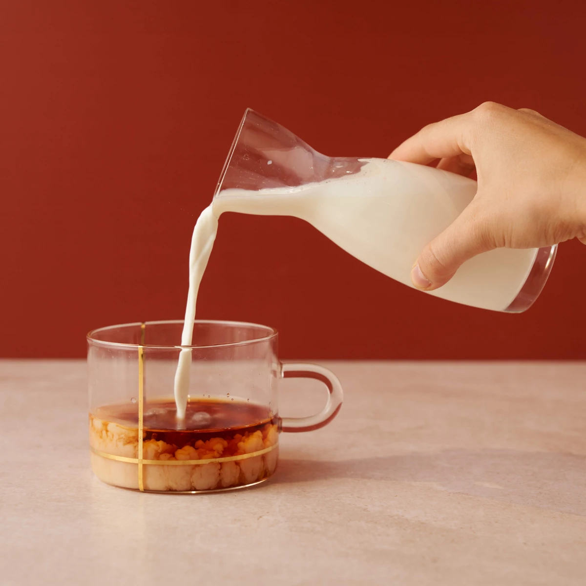 Eine Hand gießt Milch aus einer Glaskaraffe in eine Tasse Rooibos-Vanille auf einer beigen Fläche vor einem rotbraunen Hintergrund.