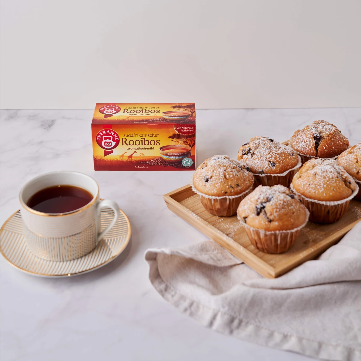 Eine gestreifte Tasse und Untertasse mit Tee stehen neben einer Schachtel Südafrikanischer Rooibos und einem Tablett mit sechs Muffins mit Puderzucker auf einer beigen Serviette, alles auf einer Marmoroberfläche angeordnet.