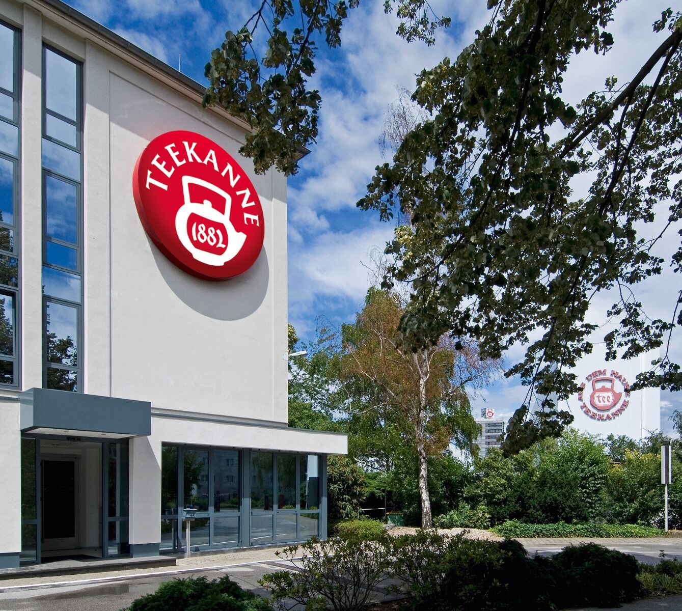 Ein weißes, modernes Gebäude mit einem großen, runden, roten Logo an der Seite. Das Logo zeigt ein weißes Teesieb mit der Zahl "(1882)" und dem Wort "TEEKANNE" in einem Kreis. Bäume und Sträucher umgeben das Gebäude.