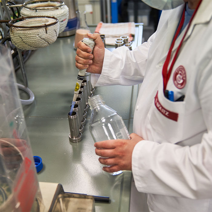 Während einer Betriebsbesichtigung verwendet eine Person im Laborkittel eine Pipette, um Flüssigkeit in den Metall-Testhalter zu geben und hält dabei eine klare Flasche im Laborumfeld.