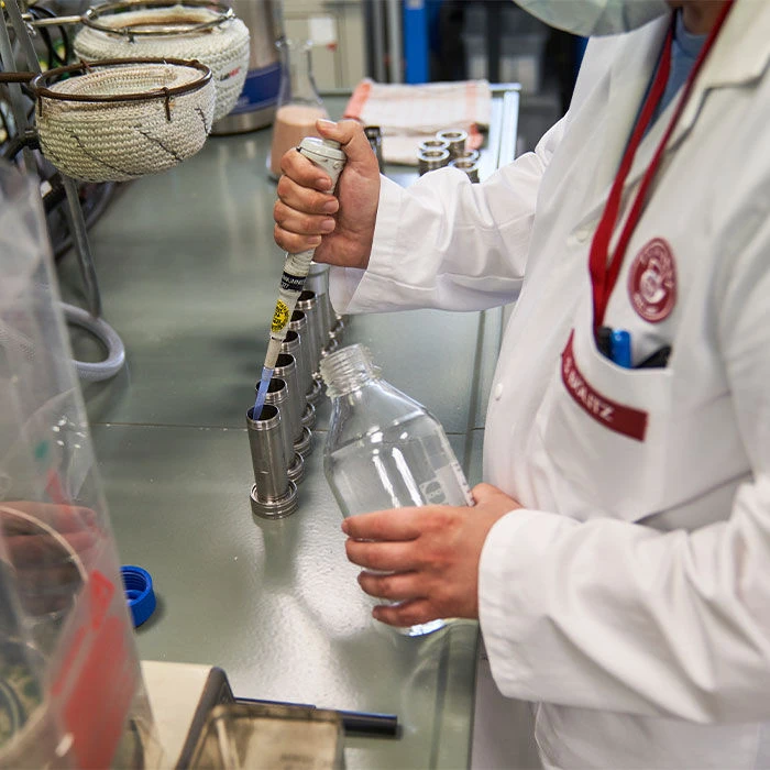 Während einer Betriebsbesichtigung verwendet eine Person im Laborkittel eine Pipette, um Flüssigkeit in den Metall-Testhalter zu geben und hält dabei eine klare Flasche im Laborumfeld.