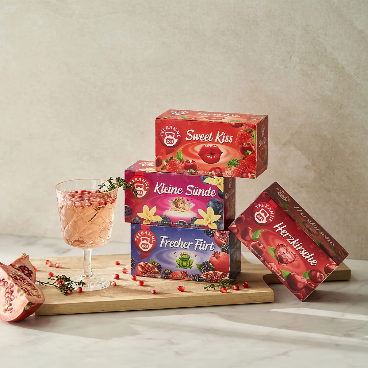 Auf einem Holzbrett sind vier gestapelte Schachteln Verführerische Früchtetees Mix & Match abgebildet, daneben ein Glas Eistee mit Granatapfelkernen und Kräutern, daneben ein halbierter Granatapfel.