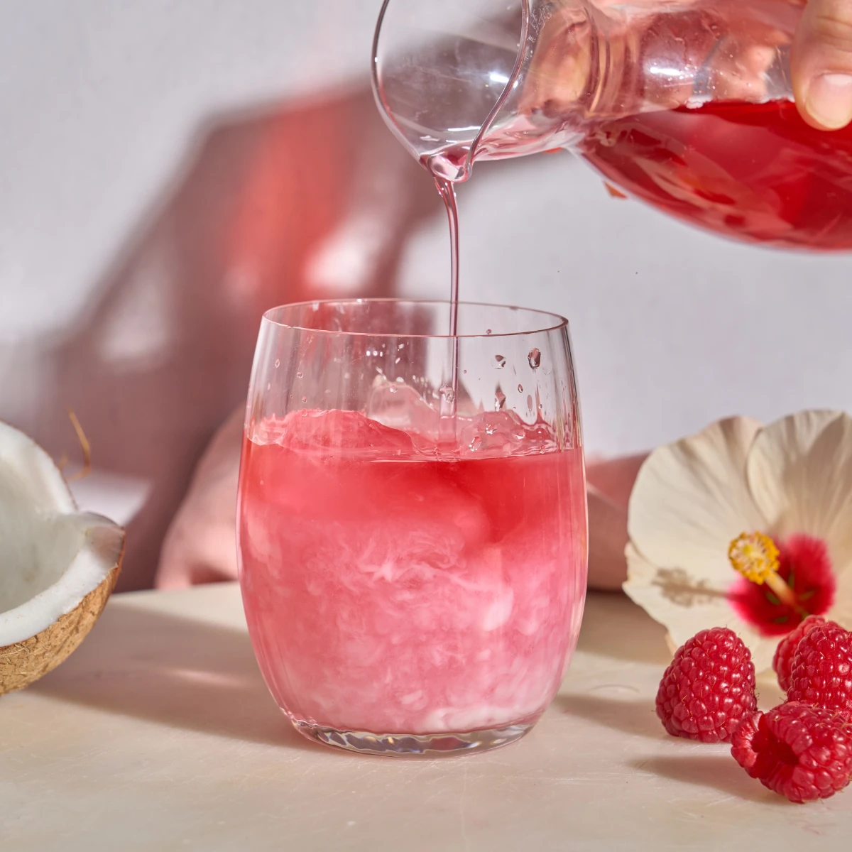 Eine Hand gießt aus einem Krug rosa Frio Kokosnuss-Hibiskus in ein Glas mit wirbelndem Rosa und Weiß, begleitet von einer halbierten Kokosnuss, frischen Himbeeren und einer blassrosa Hibiskusblüte.