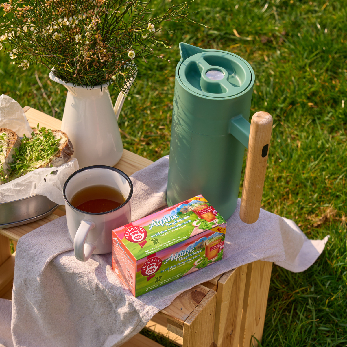 Zu einem Picknick auf der Wiese gehören eine grüne Thermoskanne, eine weiße Tasse Tee, eine Schachtel Alpine Bergkräuter, eine Blumenvase und ein Sandwich auf einer Holzkiste mit einem Tuch.