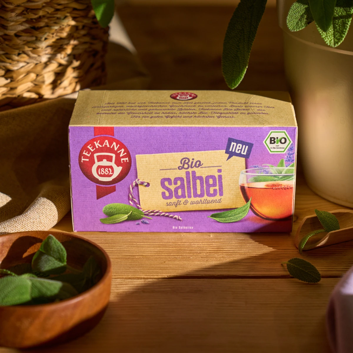 Eine Schachtel Bio-Salbei-Kräutertee steht auf einem Holztisch, umgeben von frischen Salbeiblättern, einem Weidenkorb und einer Tasse Tee im Hintergrund. Das Sonnenlicht wirft warme Schatten auf die Szene.