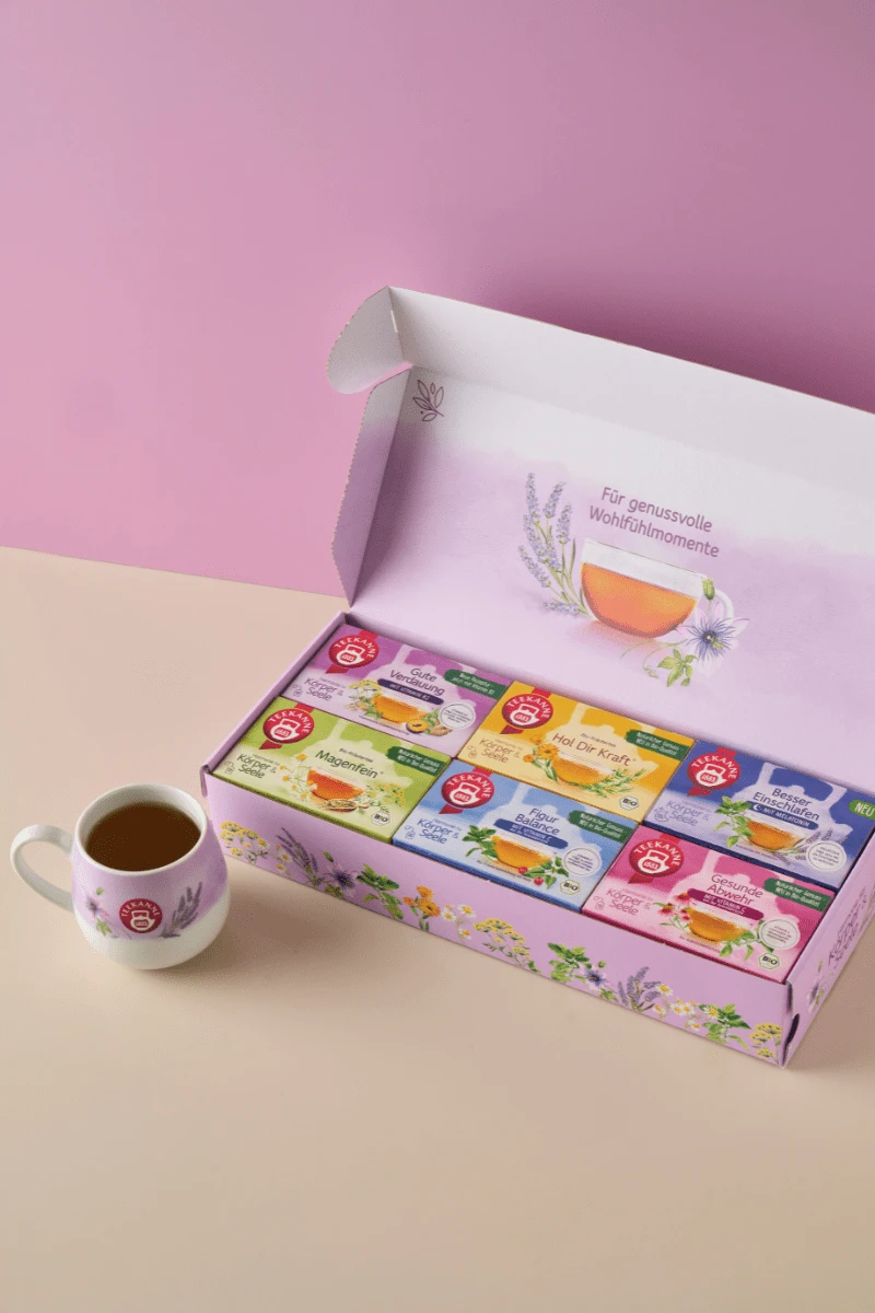Eine pastellfarbene Szene mit der Harmonie für Körper & Seele Geschenkbox - eine geöffnete Schachtel mit verschiedenen Kräuterteebeuteln mit floralen Motiven in zartem Lila und Grün - auf einer beige-rosa Oberfläche neben einer weißen Teetasse.