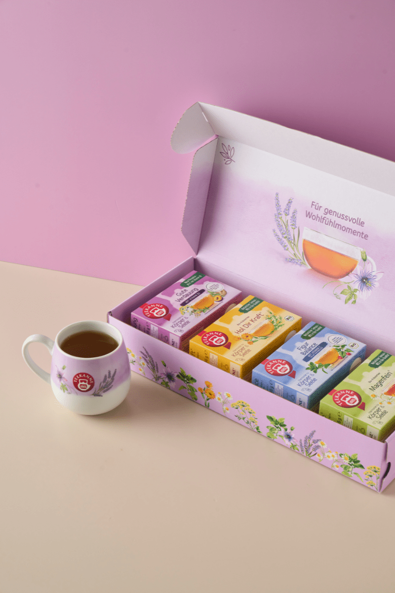 Die Harmonie für Körper & Seele Geschenkbox, eine pastellrosa Box mit Blumenmuster, lässt sich öffnen und zeigt eine Auswahl an Kräutertees. Daneben steht ein weißer, geblümter Becher, der mit Tee gefüllt ist, vor einem rosa-beigen Hintergrund.
