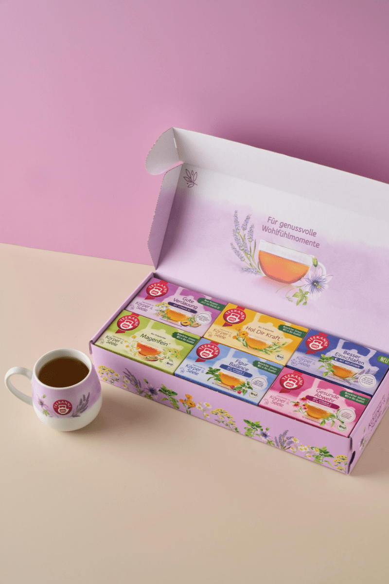 Eine pastellfarbene Szene mit der Harmonie für Körper & Seele Geschenkbox - eine geöffnete Schachtel mit verschiedenen Kräuterteebeuteln mit floralen Motiven in zartem Lila und Grün - auf einer beige-rosa Oberfläche neben einer weißen Teetasse.