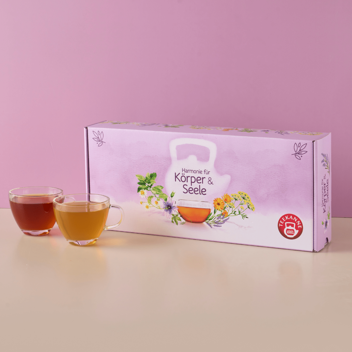 Eine pastellrosa Harmonie für Körper & Seele Geschenkbox steht neben zwei Glastassen mit Tee - einer bernsteinfarbenen und einer hellgelben - vor einem rosa Hintergrund.